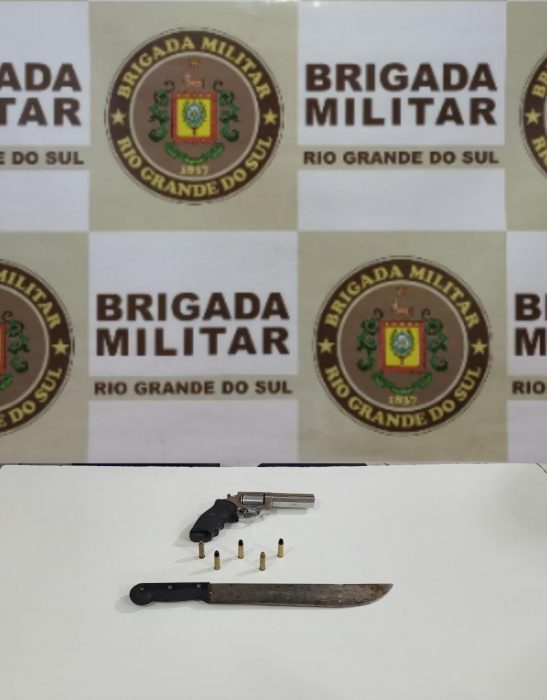 foto colorida de arma e facão apreendidos em cima de mesa branca com fundo com logo da Brigada Militar do RS