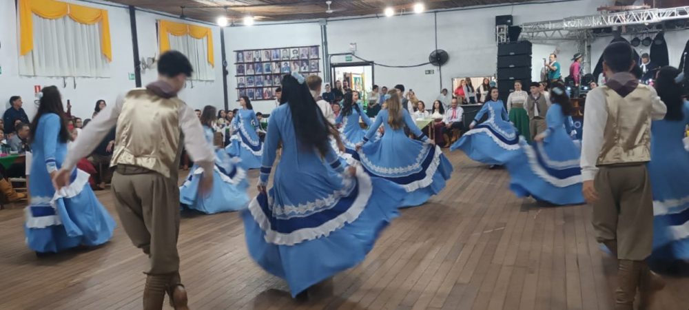 Foto colorida de apresentação de grupos de dança gaúcha no aniversário do CTG Galpão da Estância. As prendas estão de vestido azul e os peões de bombacha marrom com colete dourado.