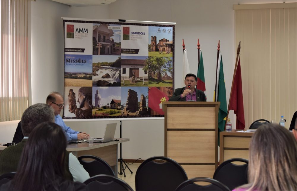 Foto colorida de reunião da AMM com um homem em púlpito, ao seu lado está um banner com imagens dos municípios
