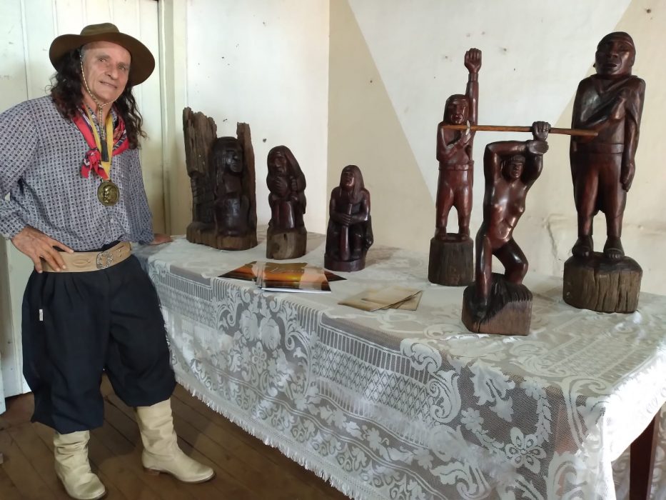 Foto colorida do artesão Marquito Moraes, um homem branco com cabelo comprido e pilchado, com camisa, lenço, bombacha, bota e chapéu. Ao seu lado diversas esculturas em uma mesa com toalha branca.